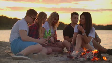 Five-young-people-are-sitting-in-shorts-and-T-shirts-around-bonfire-on-the-sand-beach.-They-are-talking-to-each-other-and-drinking-beer-at-sunset.